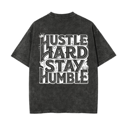 A dark gray mineral-wash t-shirt with a large white graphic text print reading 'HUSTLE HARD STAY HUMBLE' on the back, shown against a plain white background.