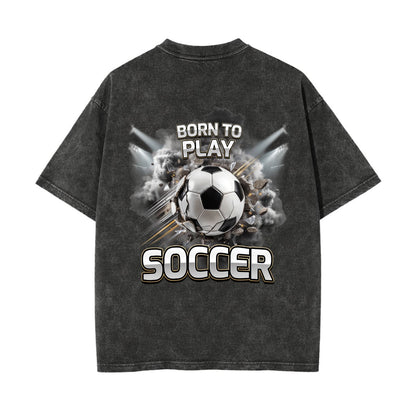 A dark gray acid-wash t-shirt shown from the back, featuring a large graphic print of a soccer ball bursting through smoke with the text 'BORN TO PLAY' above and 'SOCCER' below.