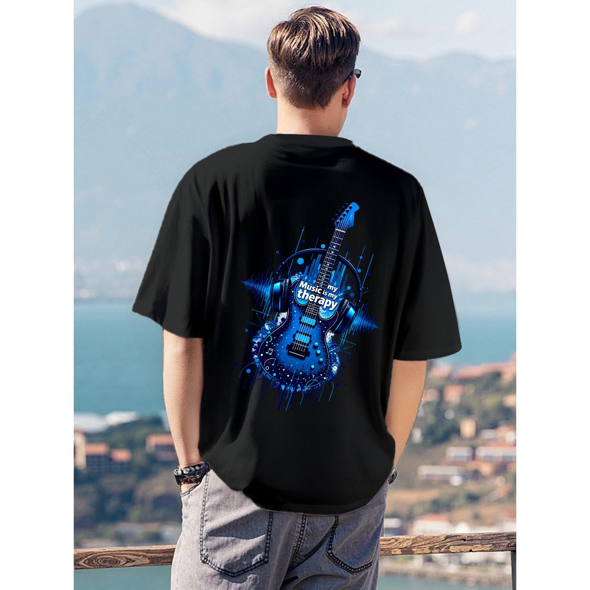 A man viewed from behind wearing a black t-shirt with a large blue electric guitar graphic on the back, standing outdoors with a scenic ocean and mountain background.