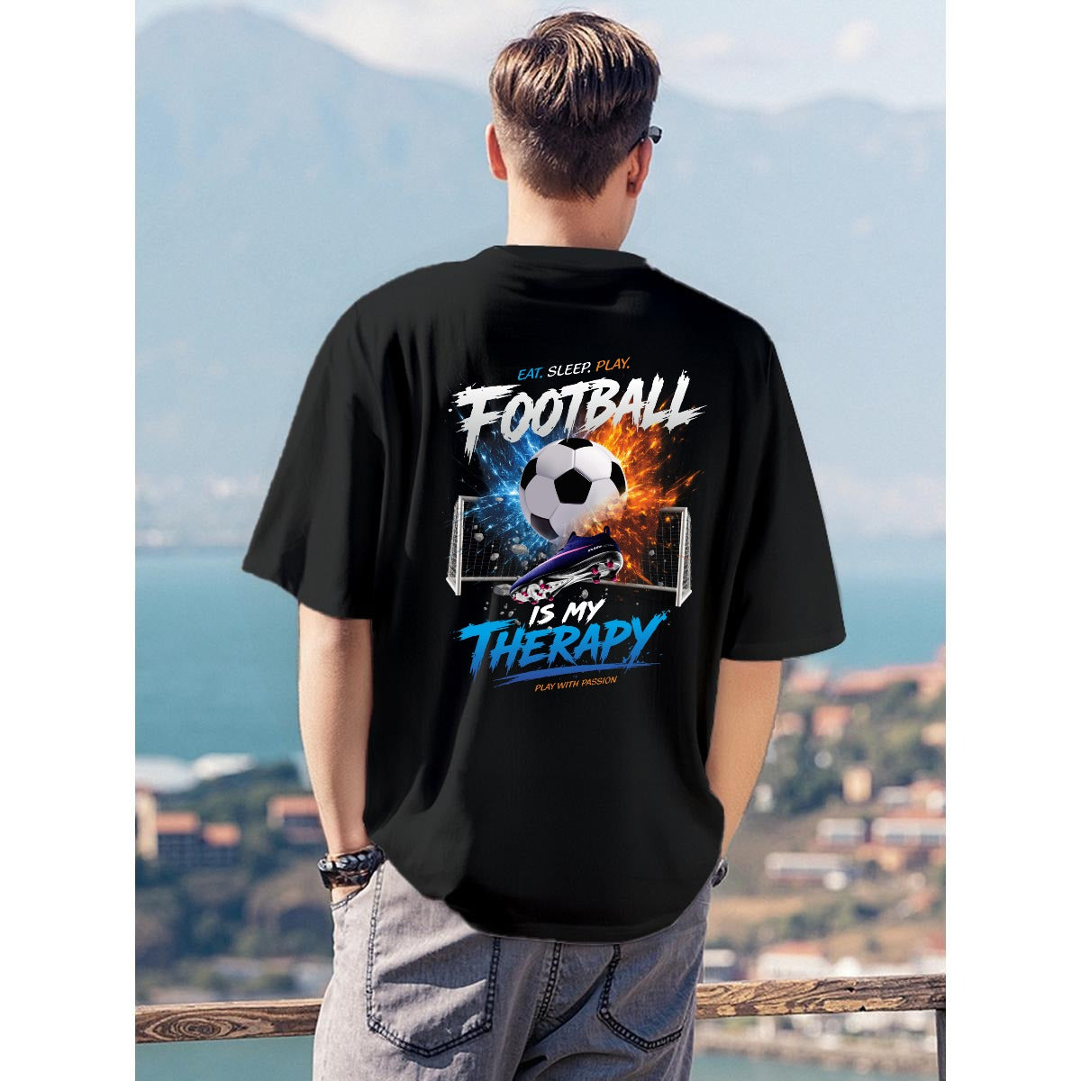 A man viewed from behind wearing a black t-shirt with a large graphic print on the back featuring a soccer ball, goals, and the text 'FOOTBALL IS MY THERAPY'. The background shows a scenic coastal view with mountains and sea.