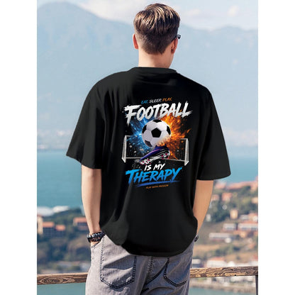 A man viewed from behind wearing a black t-shirt with a large graphic print on the back featuring a soccer ball, goals, and the text 'FOOTBALL IS MY THERAPY'. The background shows a scenic coastal view with mountains and sea.
