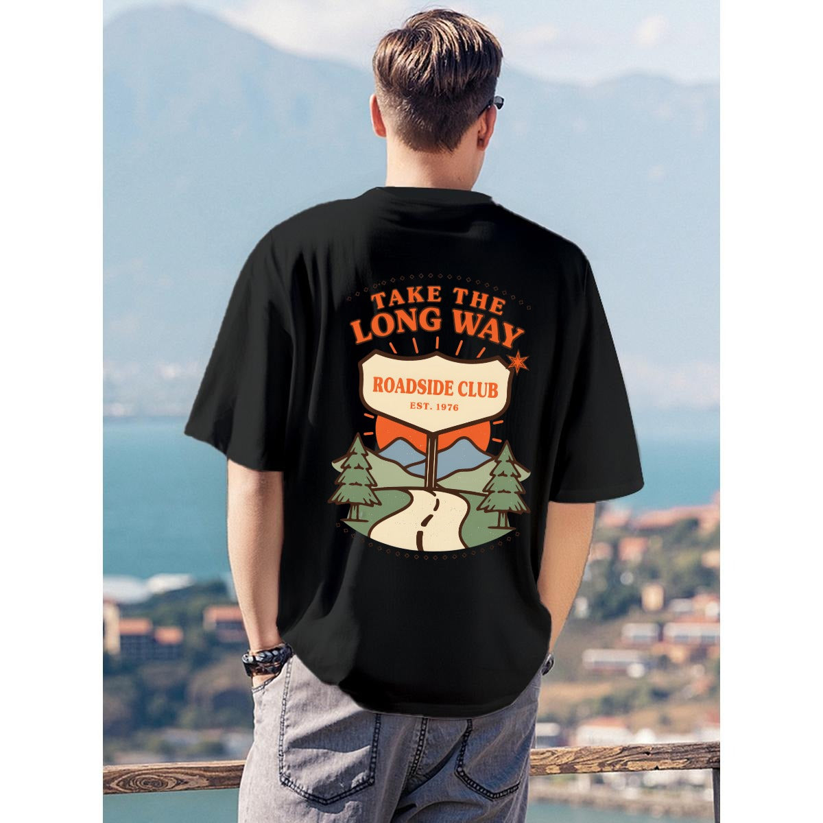 A man viewed from behind wearing a black graphic t-shirt with a retro-style 'Roadside Club' design on the back, set against a scenic coastal background.