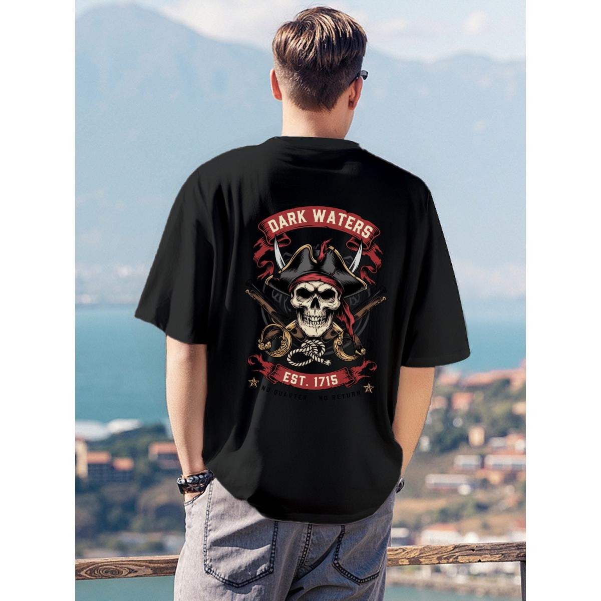 A man viewed from behind wearing a black t-shirt with a large pirate-themed graphic on the back, standing outdoors with a scenic coastal background.