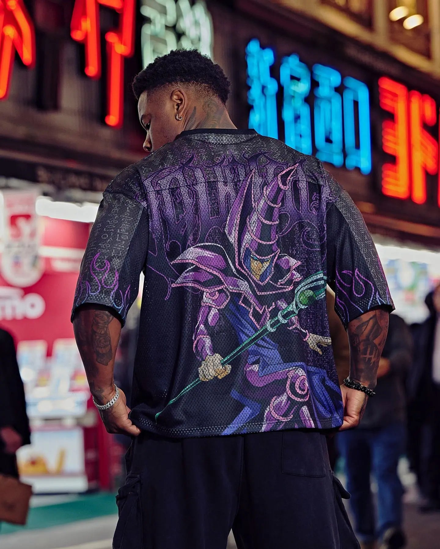Person wearing a black and purple oversized anime t shirt for men with a mystical sorcerer anime design, standing in front of neon signs.