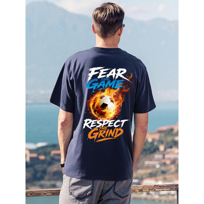 A man viewed from behind wearing a navy blue t-shirt with a large graphic on the back. The graphic features a soccer ball engulfed in flames with the text 'FEAR THE GAME' in blue and 'RESPECT THE GRIND' in orange and white.
